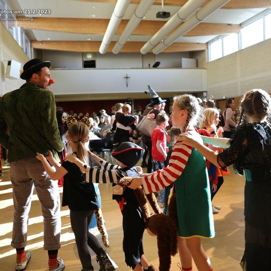 Eine Gruppe von Menschen ist in einem Saal versammelt, einige halten Händchen, ein Mann trägt Hut und Clownsnase. Der Raum hat Holzbalken und eine Bühne mit einem Projektor.