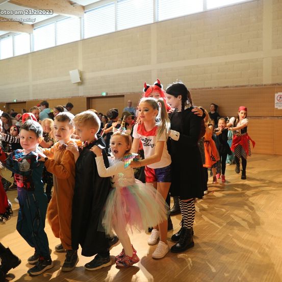 Kinder in Kostümen tanzen in einem großen Raum, mit einem Mädchen in einem rosa Tutu und einem anderen in einem weißen Shirt vorne stehend.