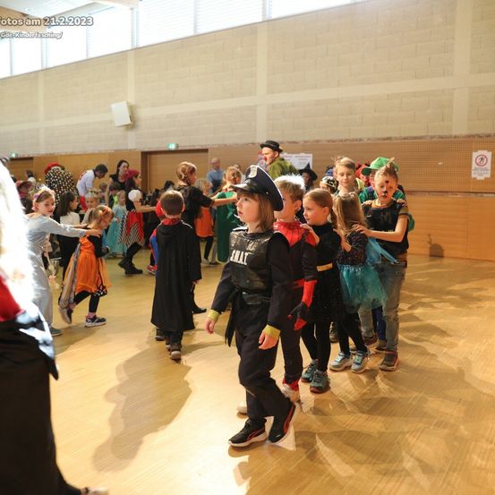 Kinder in Kostümen tanzen in einer Halle, einige tragen Hüte, Masken und Schuhe. Eine Person steht im Vordergrund. Ein Wasserzeichen ist oben links.