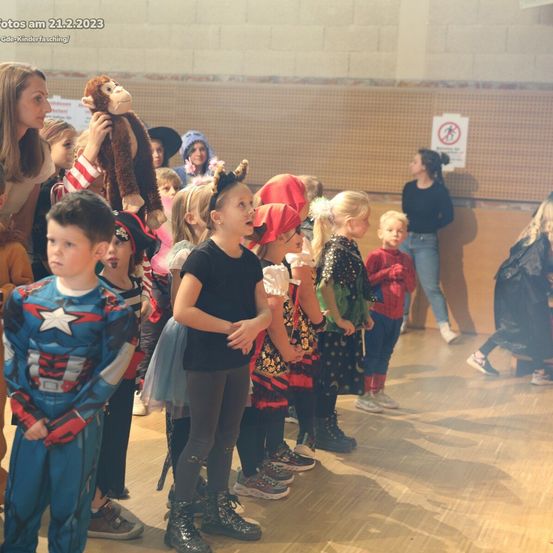 Kinder in Kostümen versammeln sich in einer Halle, einige in Superheldenkostümen, eines mit einer Affenpuppe, eine Erwachsene in der Nähe, ein Schild an der Wand und ein Schatten auf dem Boden.