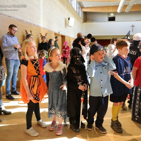 Mehrere Kinder in Kostümen stehen in einem Raum, wahrscheinlich bei einer Schulveranstaltung. Zwei Mädchen in Schmetterlings- und Prinzessinenkostümen stehen zusammen, und ein Junge in einem Darth-Vader-Kostüm hält einen Gehstock. Dahinter sind ein Junge in einem Polizeiuniform und ein weiterer in einem Piratenkostüm zu sehen.
