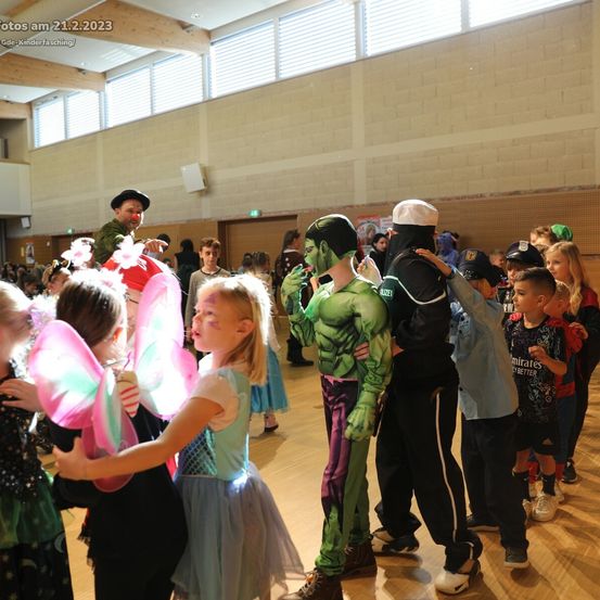 Eine Gruppe von Kindern in verschiedenen Kostümen versammelt sich in einer Halle. Einige tragen Feenflügel, andere sind Superhelden. Ein Erwachsener in Clown-Kleidung ist dabei, während eine weitere Person in einem Hulk-Kostüm ein Kind ansieht. Die Szene spielt drinnen.