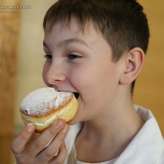 Ein junger Junge isst einen pulverisierten Donut mit dem Mund. Er hält den Donut in beiden Händen und beißt hinein. Er trägt ein weißes Hemd.