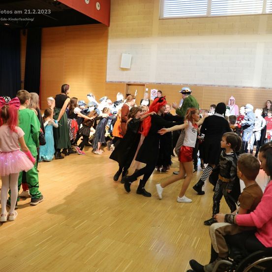 In einem Saal tanzen viele Kinder in verschiedenen Kostümen. Der Boden ist aus Holz und im Hintergrund befindet sich eine Bühne. Einige Erwachsene stehen herum und beobachten.