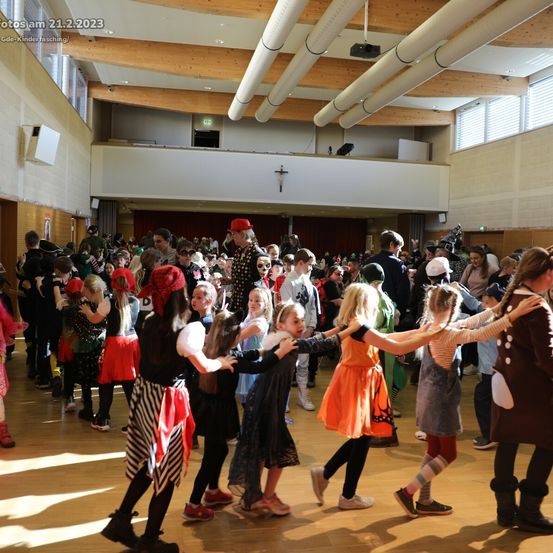 Eine große Gruppe von Kindern in Kostümen tanzt in einem Saal, mit einer Person in der Mitte, die einen roten Hut und ein rotes Bandana trägt.