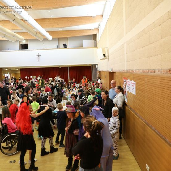 Ein überfüllter Raum mit Menschen in Kostümen, darunter ein Mädchen im Rollstuhl. Sie versammeln sich in einem, was wie eine Schulcafeteria aussieht. Einige tragen Masken, und es gibt ein Schild an der Wand.