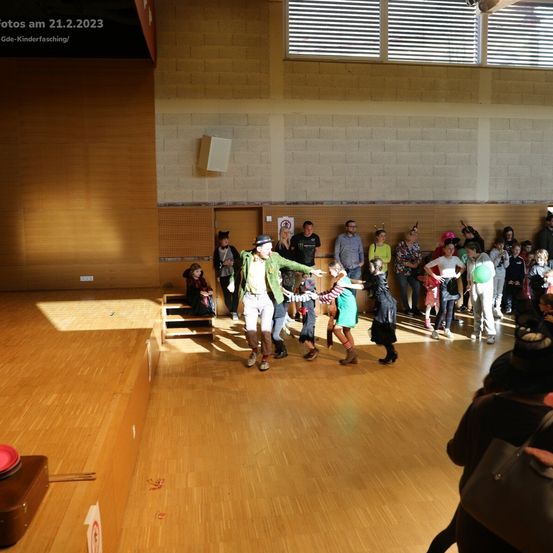 Eine Gruppe von Menschen in einem Saal, möglicherweise bei einer Schulveranstaltung, sind in verschiedenen Kostümen gekleidet. Ein Mann in einem grünen Outfit tanzt mit einem Mädchen in einem grünen Kleid. Ein roter Teller und ein Koffer liegen auf dem Boden.