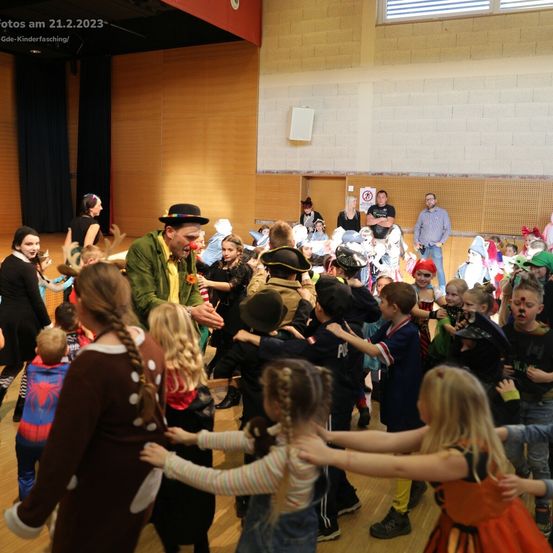 Eine Gruppe von Kindern in Kostümen tanzt in einem Gymnastiksaal um einen Clown. Der Clown trägt eine rote Nase und einen Hut. Mehrere Erwachsene beobachten von den Seitenlinien. Das Bild ist vom 21.2.2023.
