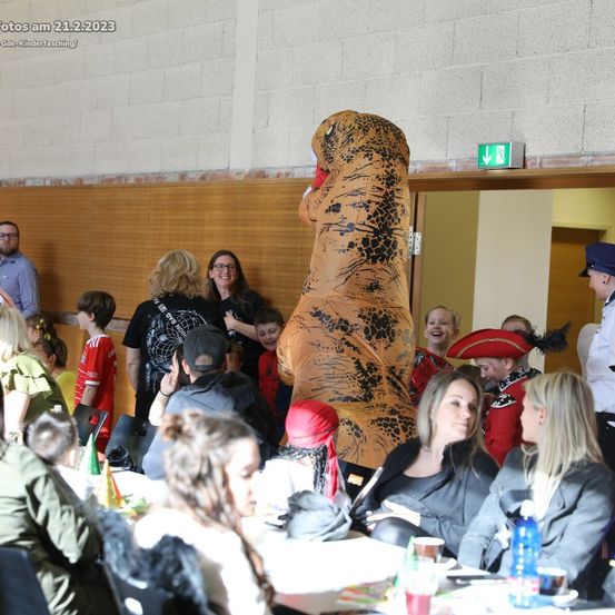 Eine Indoor-Veranstaltung mit Menschen an Tischen, einer Person in einem Dinosaurierkostüm, einer Person in einem Piratenhut und einem Notausgangsschild. Datum: 21.2.2023.