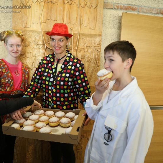 Eine Frau in einer gepunkteten Jacke hält eine Schachtel mit Donuts. Ein Junge in einem weißen Laborkittel isst einen Donut. Dahinter steht ein Mädchen in einem rosa T-Shirt und mit Sonnenbrille. Datumsstempel auf dem Bild: 21.2.2023.
