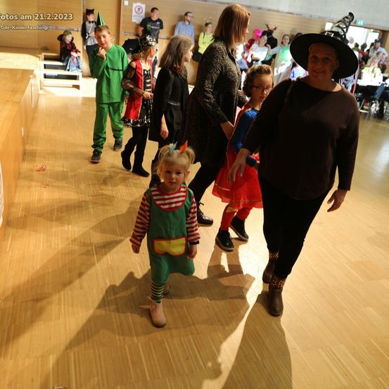 Eine Gruppe von Kindern in Kostümen geht in einem Saal, einige von Erwachsenen begleitet. Der Boden ist aus Holz, und es gibt Zuschauer im Hintergrund.
