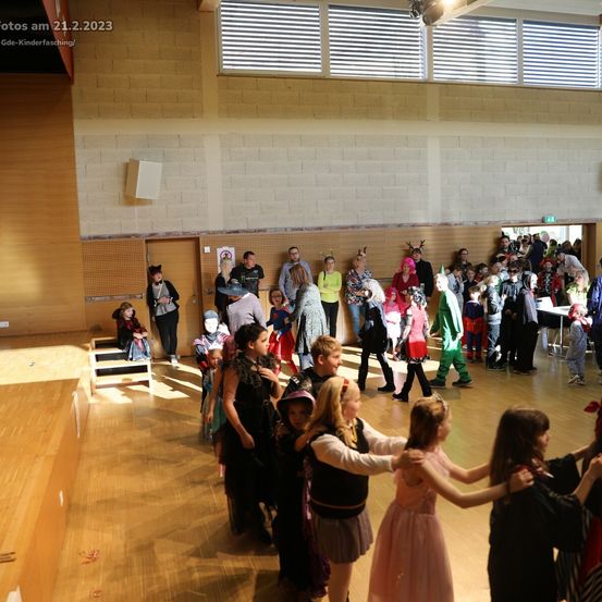 Eine Gruppe von Kindern in verschiedenen Kostümen tanzt am 21. Februar 2023 in einer Halle. Die Halle hat Holzböden, eine Bühne und weiße Ziegelwände.