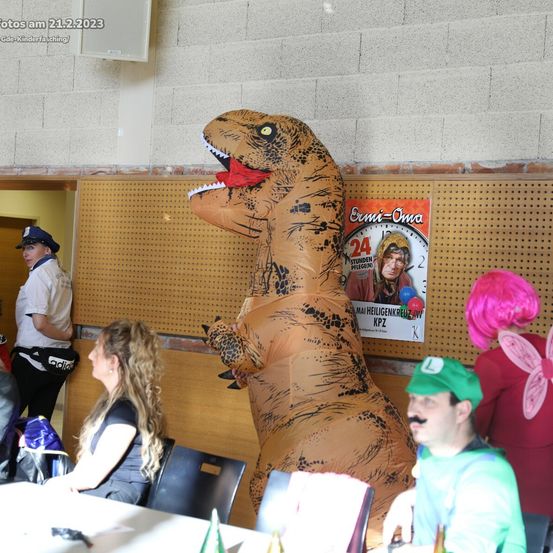 Eine Person in einem großen aufblasbaren Dinosaurierkostüm steht unter anderen in einem Raum mit einem Schild an der Wand. Eine Frau mit langen Haaren und ein Mann in einem grünen Kostüm sitzen an einem Tisch.