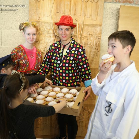 Eine Frau in einem gepunkteten Anzug steht mit einer Schachtel Donuts, umgeben von Kindern, eines isst einen Donut und ein anderes hält die Schachtel. Das Datum 21.2.2023 ist oben sichtbar.
