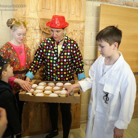 Eine erwachsene Frau in einem gepunkteten Outfit hält eine Schachtel mit Donuts, umgeben von Kindern. Ein Kind in einem weißen Laborkittel hält die Schachtel. Das Foto ist vom 21.2.2023.