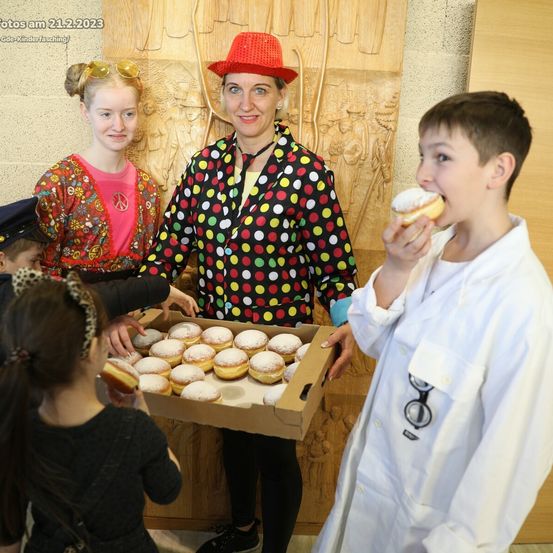 Eine Frau in einem Punktemuster-Kostüm hält eine Schachtel mit Donuts, während ein Junge in einem Laborkittel einen isst. Zwei Kinder stehen in der Nähe, und ein Mädchen trägt eine Mütze und eine Brille.