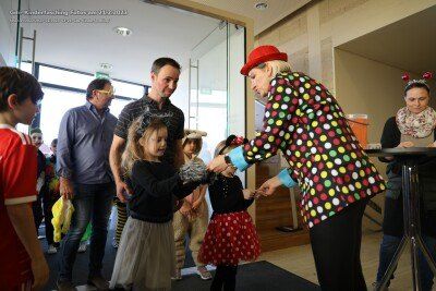 Ein Clown in einem Punkte-Kostüm überreicht einem Kind in einem roten Hut ein Geschenk. Dahinter stehen mehrere Menschen und beobachten. Der Ort scheint im Innenraum zu sein, mit einer Glaswand und einem Teppichboden.