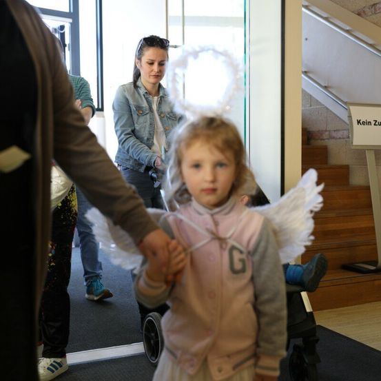 Ein kleines Mädchen, das als Engel verkleidet ist, wird von einem Erwachsenen durch eine Tür geführt, mit einer Frau und einem Schild mit der Aufschrift Kein Zu im Hintergrund.
