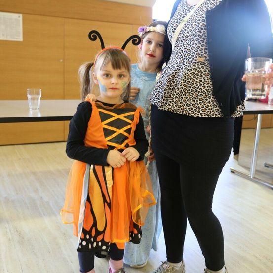 Ein junges Mädchen in einem Schmetterlingskostüm steht mit einer Frau in einem Leopardenmuster-Cardigan und einem anderen Mädchen in einem blauen Kleid in einem Raum mit Holzböden.