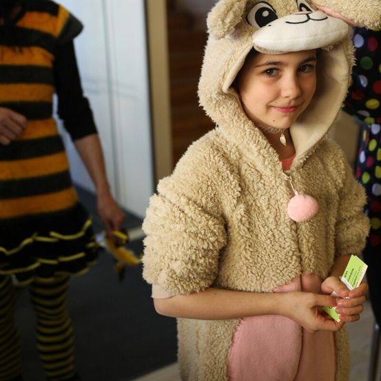 Ein kleines Mädchen in einem Schafskostüm, lächelnd und einen kleinen Gegenstand haltend, steht neben einer Person in einem Bienenkostüm.