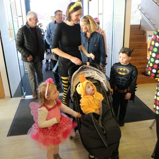 Mehrere Erwachsene und Kinder in Kostümen sind in einem Gebäude. Eine Frau hält einen Kinderwagen mit einem Baby in einem Hühnerkostüm. Ein Mädchen in einem Feenkostüm und ein Junge in einem Batman-Kostüm stehen in der Nähe.