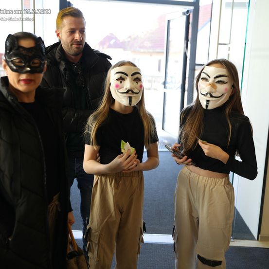 Vier Personen in Masken posieren für ein Foto in einem Gebäude. Zwei Mädchen tragen Guy-Fawkes-Masken, während die anderen Katzen- und einfache schwarze Masken tragen. Ein Mann steht hinter ihnen.
