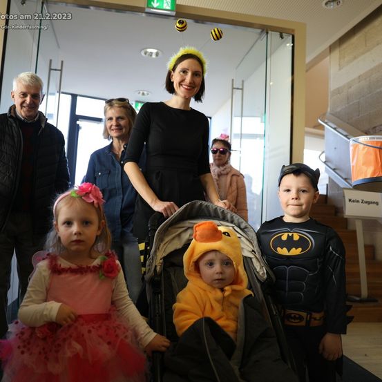 Ein Familienfoto vom 21. Februar 2023 bei einem Kinderfasching. Eine Frau in einem schwarzen Kleid steht mit einem Kinderwagen, während ein Mädchen in einem rosa Kleid und ein Junge in einem Batman-Kostüm neben ihr stehen. Hinter ihnen stehen ein Mann und eine Frau, und im Hintergrund befindet sich eine Treppe.