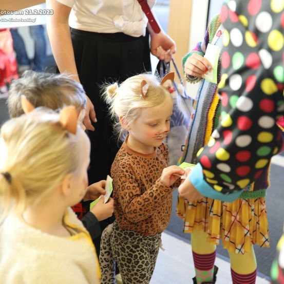 Eine Gruppe von Kindern in einem Flur, ein Mädchen mit Leopardenmuster-Kleidung und Katzenohren erhält etwas von einer anderen Person. Die anderen sind in Kostümen gekleidet. Eine Frau in einer schwarzen Rock steht im Hintergrund.