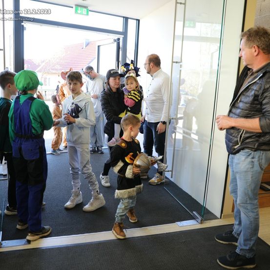 Menschen in Kostümen versammeln sich drinnen; einer hält ein Baby, der andere einen Helm. Ein Mann spricht mit einem anderen in der Nähe einer Glastür. Das Bild ist vom 21.2.2023.