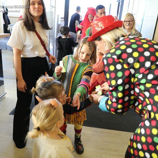 Eine Gruppe von Kindern und Erwachsenen in einem Saal. Eine Frau in einer Punktekleidung verteilt Tickets an Kinder in Kostümen.