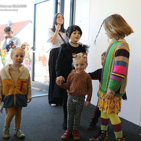 Eine Gruppe von Kindern in Kostümen steht in einem Raum, mit einer Frau dahinter. Einige tragen Katzenohren, andere haben Gesichtsbemalung. Das Datum und der Ort sind oben links angegeben.
