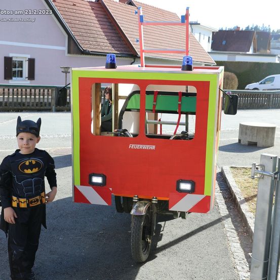 Ein Junge in einem Batman-Kostüm steht neben einem Feuerwehrauto. Das Auto hat blinkende Lichter auf dem Dach.