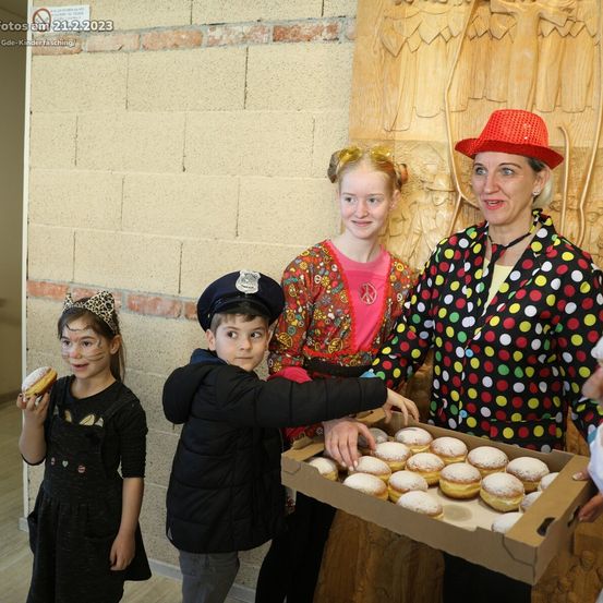 Drei Kinder und eine erwachsene Frau, gekleidet in festliche Kleidung, stehen an einer Ziegelmauer. Die Frau hält eine Schachtel mit Donuts. Die Kinder tragen Gesichtsbemalung und Kostüme. Ein Schild mit der Aufschrift 'Rauchen verboten' befindet sich an der Wand.