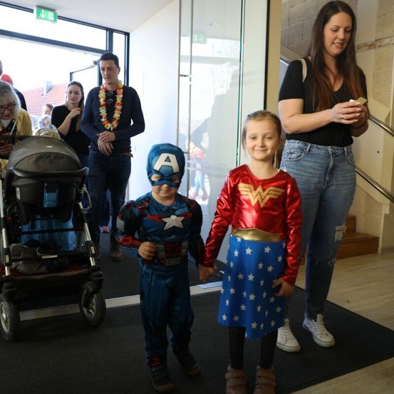 Ein junger Junge in einer Captain America-Kostüm und ein Mädchen in einem Wonder Woman-Kostüm stehen zusammen vor einem Eingang. Dahinter schauen ein Mann und eine Frau auf ihre Handys.
