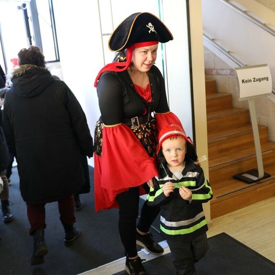 Eine Frau in einer Piratenkostüm und ein Kind in einer reflektierenden Jacke gehen durch einen Gebäudekorridor.
