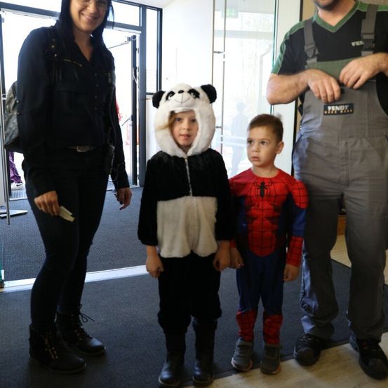 Zwei Kinder in Kostümen, eines als Panda und das andere als Spiderman, stehen zwischen einer Frau und einem Mann in Overalls in einem Flur.