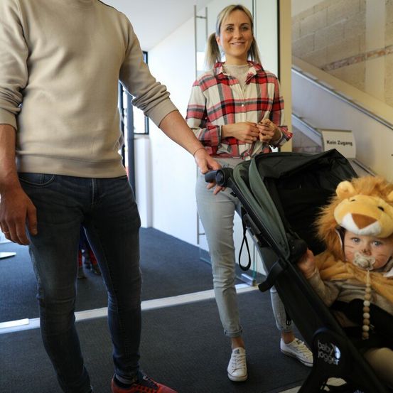 Eine Familie steht in einem Flur, wobei ein Mann die Hand einer Frau hält. Die Frau hält einen Kinderwagen mit einem Baby in einem Löwenkostüm. Der Flur hat eine weiße Wand, eine Glastür und eine Treppe.