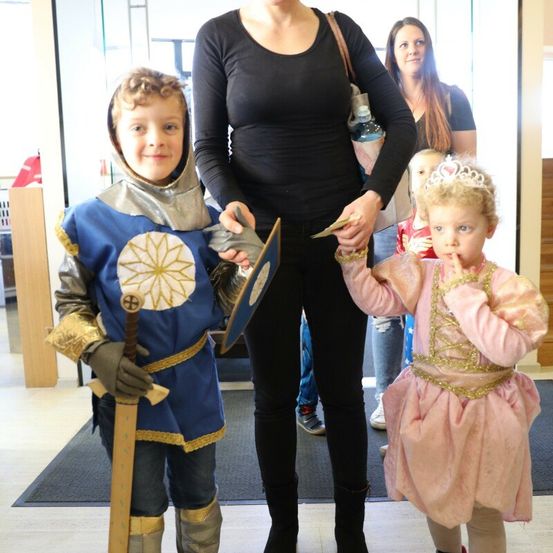 Eine Frau steht mit zwei Kindern in Kostümen, ein Junge mit einem Schwert und ein Mädchen mit einer Krone, in der Nähe eines Eingangs. Eine Frau im Hintergrund trägt ein Kind.