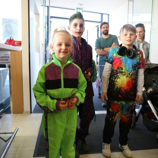 Drei Kinder stehen lächelnd zusammen in einem Flur. Ein Mädchen in Grün, ein Junge in bunten Farbspritzern und ein Junge in dunklen Farben. Hinter ihnen beobachten zwei Erwachsene. Der Flur hat Glaswände, eine Tür und einen Teppich.