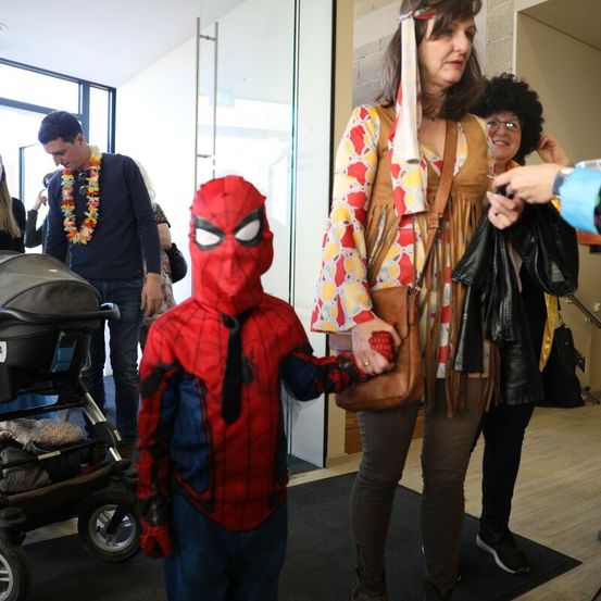 Ein junger Junge, verkleidet als Spider-Man, steht neben einer Frau in bunter Kleidung in einem Flur. In der Nähe ein Mann mit einer Blumenkette und einem Kinderwagen und eine weitere Frau mit einer Handtasche.