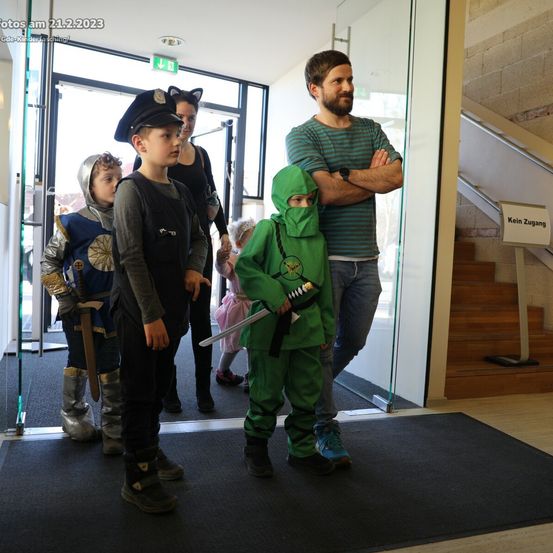 Mehrere Kinder in verschiedenen Kostümen, einschließlich Ritter, Pirat und Ninja, stehen vor einer Glastür, wobei Erwachsene beobachten. Die Umgebung scheint ein Flur mit Treppen zu sein.
