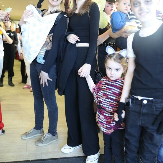 Eine Gruppe von Menschen steht in einer Reihe, zwei Frauen halten Babys auf ihren Schultern, und ein kleines Mädchen hält die Hand einer Person mit einer Mausohr-Kopfbedeckung.