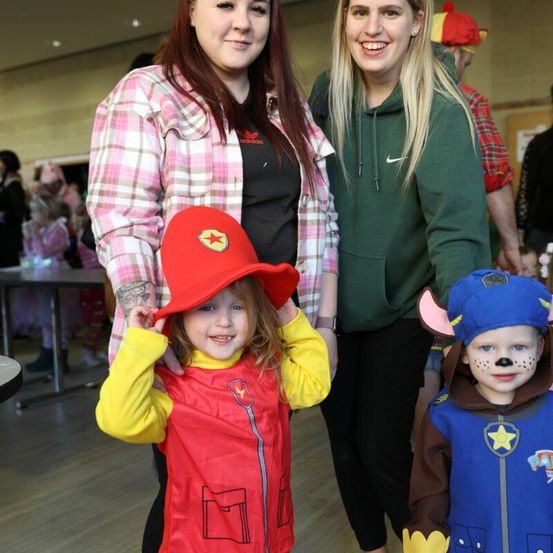 Zwei Frauen stehen neben zwei Kindern in Kostümen, eines verkleidet als Feuerwehrmann und das andere als Polizist. Sie alle lächeln und scheinen sich auf einem Kostümfest zu befinden.