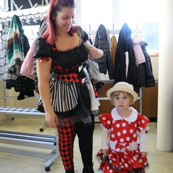 Eine Frau in einem schwarz-weißen Kostüm steht mit einem kleinen Mädchen in einem roten Punktekleid in einer Umkleidekabine mit an den Stangen hängenden Kleidern.
