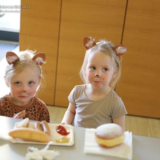 Zwei Mädchen mit Katzenohren und Katzen-Make-up sitzen an einem Tisch, eines mit einem Hotdog und das andere mit einem Donut. Datum: 21.2.2023.