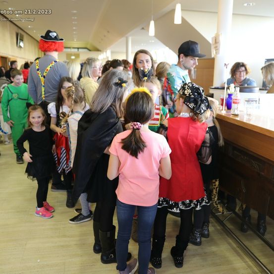 Eine Gruppe von Kindern und Erwachsenen steht in einer Schlange an einer Theke, einige in Kostümen gekleidet. Ein Erwachsener in Clown-Kostüm steht hinter ihnen.