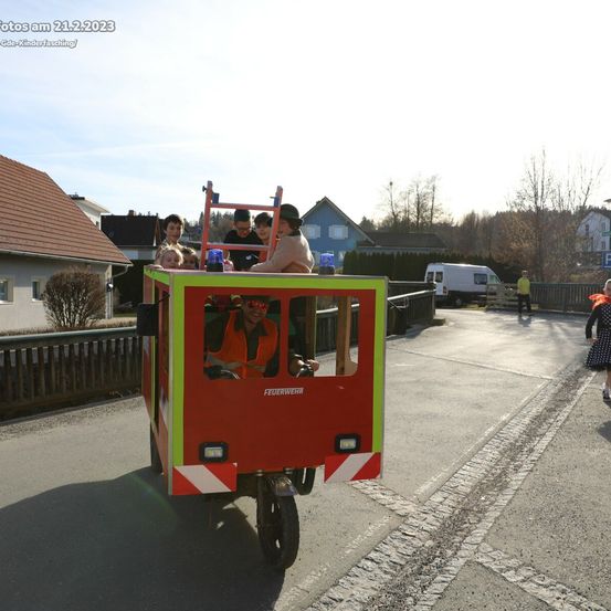 Ein Feuerwehrauto mit Kindern fährt auf einer Straße, mit Häusern und Menschen im Hintergrund. Das Foto ist vom 21.2.2023.