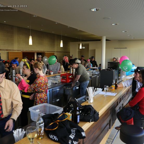 Eine Menschenmenge in farbenfrohen Perücken und Kostümen versammelt sich um eine Bar in einem Restaurant. Die Bar ist gut bestückt mit Flaschen, Gläsern und Luftballons. Einige Leute sitzen auf Hockern, andere stehen. Der Raum ist hell beleuchtet.