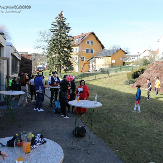 Eine Gruppe von Menschen versammelt sich um Tische im Freien, einige in Kostümen. In der Nähe spielt ein Kind auf dem Rasen. Im Hintergrund sind Häuser und ein Spielplatz zu sehen. Das Datum ist als 21. Februar 2023 gestempelt.