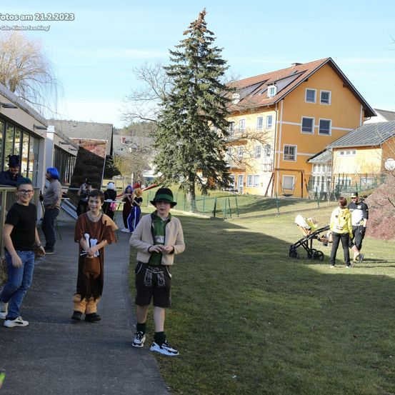 Eine Gruppe von Menschen in Kostümen geht auf einem Bürgersteig, mit einem gelben Haus und einer Schaukel im Hintergrund.